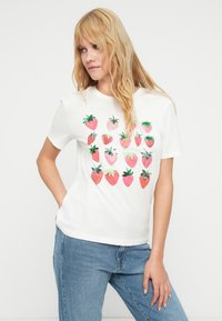 Λευκό βαμβακερό t-shirt με γραφικό τύπωμα κόκκινων φραουλών με πράσινα φύλλα. Κοντά μανίκια και χαλαρή εφαρμογή για μεγαλύτερη άνεση.