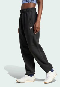 Černé tepláky s elastickým pasem, bočními kapsami a logem Adidas. Stylizované s bílými teniskami a námořnickými ponožkami.