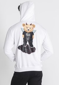 Biela mikina s grafikou na chrbte zobrazuje medveďa s krídlami a diablovými rožkami, ktorý nosí čierne tričko s textom, zasadená do dizajnu oblakov.
