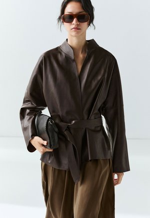 Femme portant une veste en cuir marron ceinturée et des lunettes de soleil, tenant une pochette noire sur un fond clair uni.