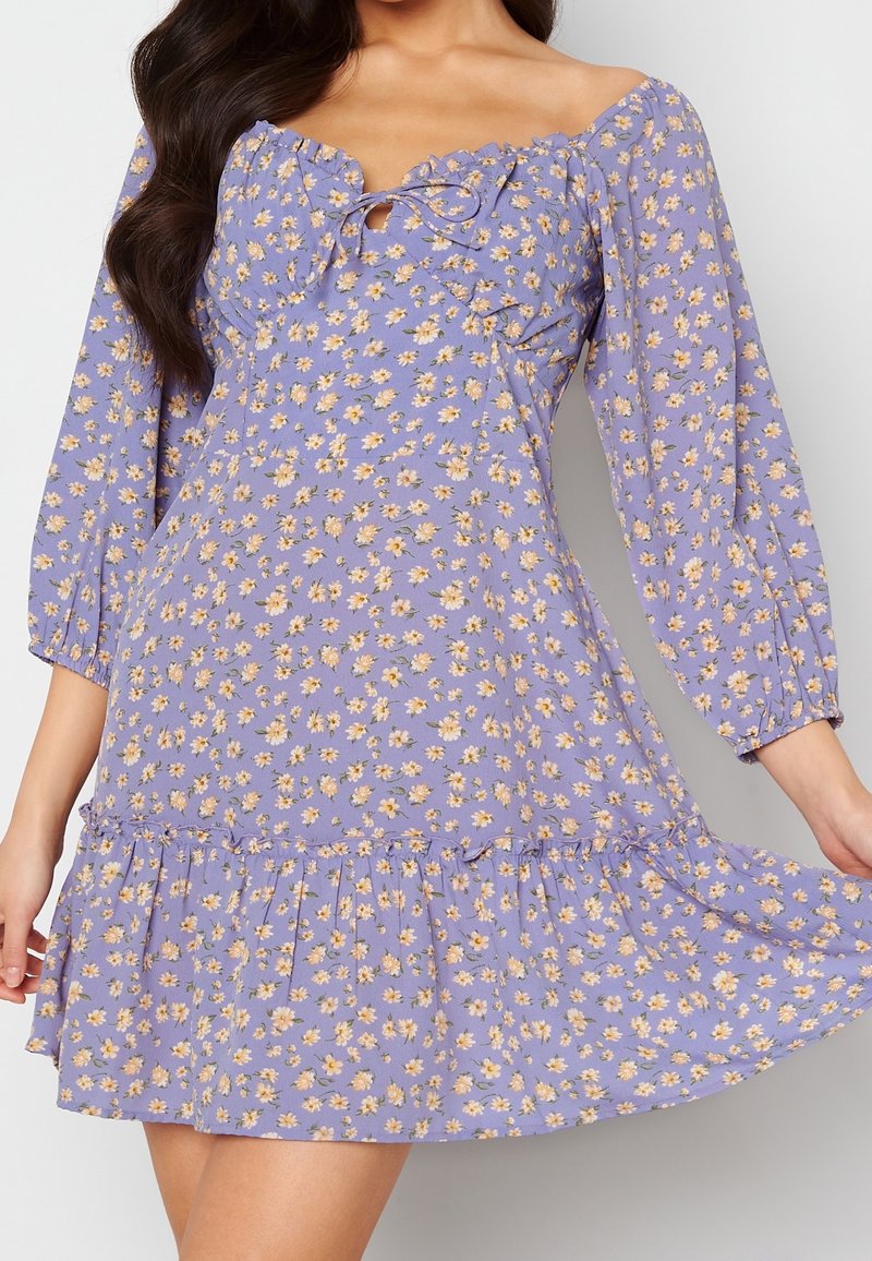 Langen, puffärmeliger lila Blumenkleid mit rüschiertem Ausschnitt und gestuftem Saum. Es hat kleine gelbe Blumen und eine Schnürung auf der Brust.