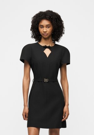 Femme aux cheveux bouclés portant une robe noire ajustée à manches courtes, avec un nœud au niveau de l'encolure et une ceinture fine à la taille.