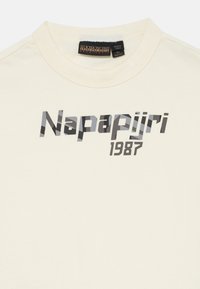 Kremfarget T-skjorte med teksten "Napapijri 1987" i svart og grått mønster på brystet og en svart nakkelapp.