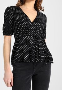 Svart blus med prickar som har en djup V-ringning, puffärmar och en skräddarsydd midja med en knytning. Mjuk, lätt tyg.