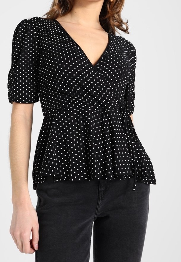 Svart blus med prickar som har en djup V-ringning, puffärmar och en skräddarsydd midja med en knytning. Mjuk, lätt tyg.