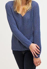 Frau trägt einen strukturierten, blauen Pullover mit V-Ausschnitt und langen Ärmeln sowie dunkle Jeans, wobei ein Arm am Handgelenk über den anderen verschränkt ist.