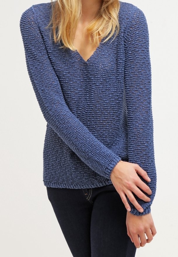 Frau trägt einen strukturierten, blauen Pullover mit V-Ausschnitt und langen Ärmeln sowie dunkle Jeans, wobei ein Arm am Handgelenk über den anderen verschränkt ist.