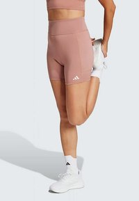 Roosa spordilühikesed püksid, millel on kõrge vöökoht, valmistatud venivast kangast. Vasakul jalal on valge Adidas'i logo. Mudel kannab valgeid jalatseid.