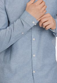 Camisa azul clara, abotoada, feita de um tecido suave, com colarinho tradicional, mangas longas com punhos e seis botões na frente.