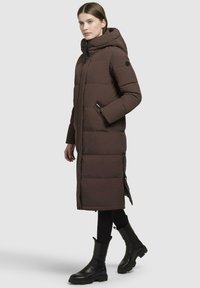 Cappotto imbottito marrone con cappuccio, dotato di chiusura con zip e bottoni, tasche laterali e design lungo, abbinato a stivaletti neri alla caviglia.