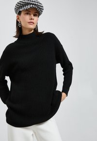 Schwarzer Strickpullover mit hohem Kragen, lockerer Passform und strukturiertem Rippenmuster. Kombiniert mit weißen Hosen und einer Hahnentrittkappe.