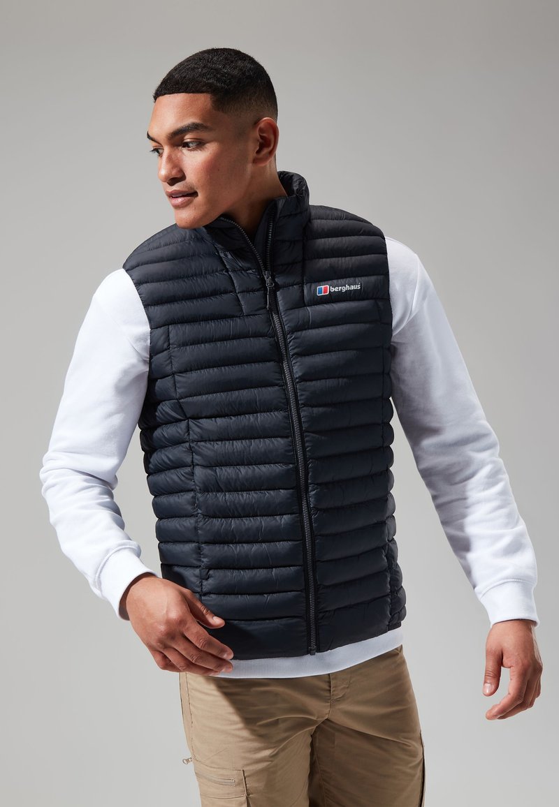 Zwarte geïsoleerde puffer vest met rits aan de voorkant, voorzien van een hoge kraag en een logopatch. Daaronder is een witte longsleeve zichtbaar.