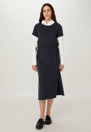 Marineblaues knielanges Kleid mit kurzen Ärmeln, Kordelzug in der Taille, seitlichem Schlitz, über einem weißen Kragenhemd getragen und mit schwarzen Schuhen kombiniert.