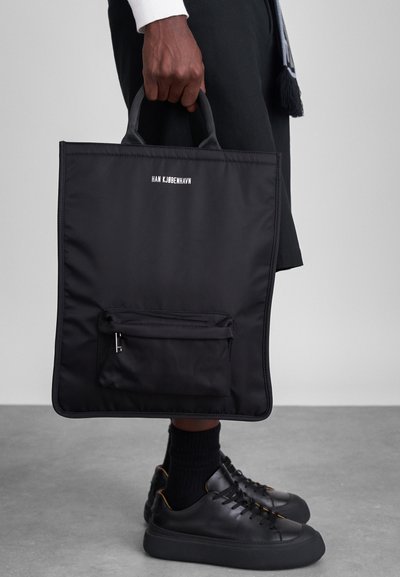 Saco de tecido preto com duas alças, apresentando um pequeno bolso frontal com fecho e o texto "HAN KJØBENHAVN" impresso na frente.