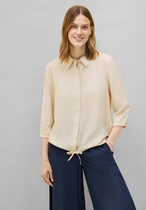 Blouse beige clair à boutons avec un col, des manches trois-quarts, un tissu texturé et un détail à nouer en bas, associée à un pantalon marine.