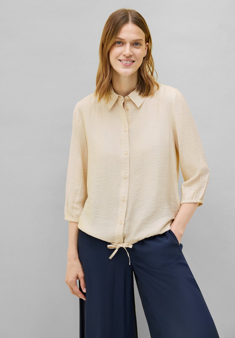 Lichtbeige blouse met knoopsluiting en kraag, driekwart mouwen, gestructureerde stof en een strikdetail aan de onderkant, gecombineerd met een marineblauwe broek.
