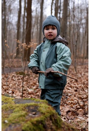 Dítě oblečené v nepromokavé bundě se dvěma odstíny – tyrkysovou a tmavě šedou, s šedou čepicí, držící hůl v lese pokrytém spadlým listím.