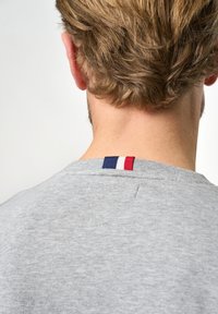 Grå hettegenser med ribbestrikket hals, med røde, hvite og blå striper på bakstykket. Myk stoff og enkelt design.