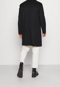 Sort, lang frakke med lige snit, kombineret med cremefarvede bukser og sorte ankelstøvler. Stoffet virker glat med få detaljer.