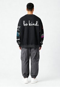 Črna dukserica z napisom "bodite prijazni." barvitimi vezeninami, v kombinaciji z sivimi cargo hlačami in belimi športnimi copati. Ohlapen kroj, dolgi rokavi.