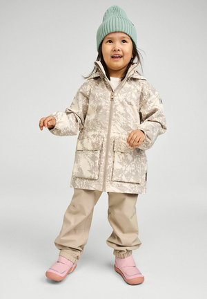 Child standing wearing beige patterned jacket, beige pants, pink boots, and light green knit hat against gray background.