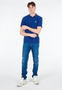 Polo shirt blu con colletto e logo, abbinato a jeans blu elasticizzati e sneakers grigie chiare, su uno sfondo bianco semplice.