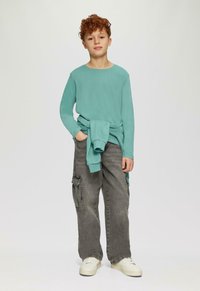 Chłopiec z kręconymi rudymi włosami, ubrany w turkusową koszulę z długim rękawem, szare krótkie spodenki cargo, białe trampki oraz turkusowy sweter zawiązany wokół talii.