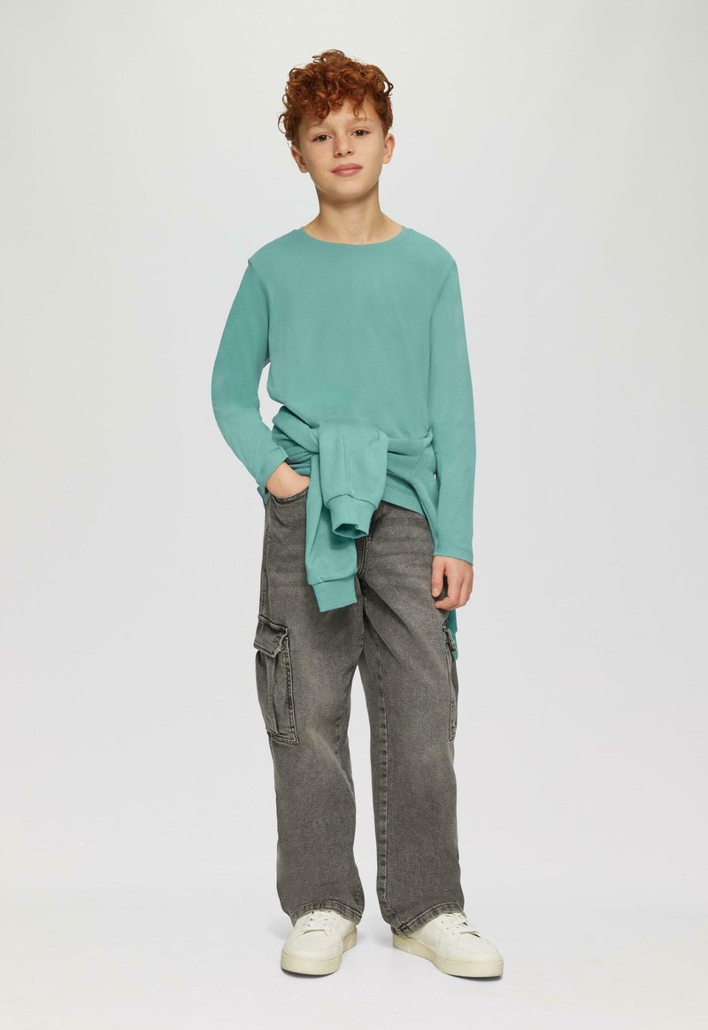 Chłopiec z kręconymi rudymi włosami, ubrany w turkusową koszulę z długim rękawem, szare krótkie spodenki cargo, białe trampki oraz turkusowy sweter zawiązany wokół talii.