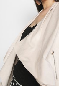 Béžová přehnutá bunda z měkké tkaniny s velkým předním klopou a postranní kapsou na zip, vrstvená přes černý žebrovaný top.