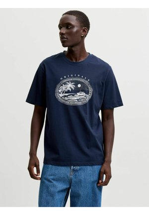 Jeune homme portant un t-shirt bleu marine avec un graphisme blanc d'île tropicale et de bateau, associé à un jean bleu, debout devant un fond uni.