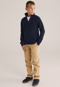 Marinblå stickad zip-up tröja med ribbad textur, ihop med beige cargobyxor och marinblå sneakers. Klädseln har en avslappnad, lager-på-lager-look.