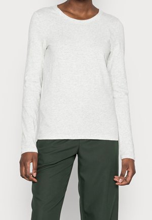 Person wearing a light gray long-sleeve shirt and dark green pants, standing against a plain background.