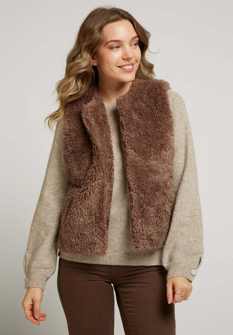 Un gilet en fausse fourrure marron superposé sur une chemise à manches longues beige clair. Le gilet présente une finition douce et texturée avec un design à l'avant ouvert.