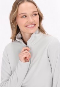 Lys grå fleecepullover med høj krave og halvlukning. Glat tekstur. Modellen holder lynlåsen med den ene hånd foran en ensfarvet baggrund.