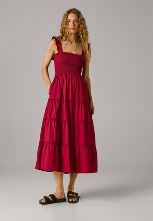 Žena s dlhými kučeravými vlasmi, ktorá má na sebe červené vrstvené midi šaty bez rukávov a čierne sandále s cvokmi, stojaca pred jednoliatym sivým pozadím.