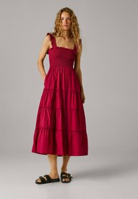 Donna con lunghi capelli ricci che indossa un vestito midi rosso a più strati senza maniche e sandali neri con borchie, in piedi davanti a uno sfondo grigio semplice.