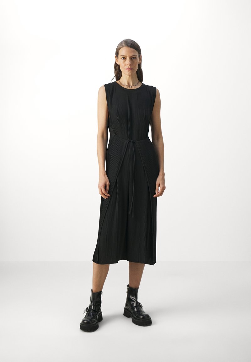 Robe sans manches noire avec une taille cintrée, présentant une texture côtelée verticale et des fentes latérales. Portée avec des bottes de combat noires.