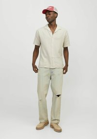 Camisa de manga corta a cuadros en color crema y verde, combinada con jeans claros rasgados y zapatos beiges. Una gorra roja y blanca completa el atuendo.