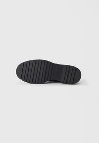 Semelle de chaussure noire avec des rainures horizontales et une zone de logo rectangulaire au centre, affichée sur un fond blanc.