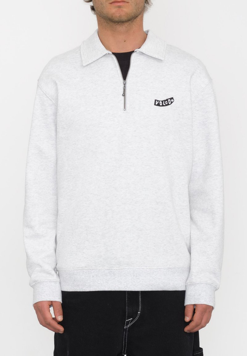 Sudadera gris claro con cuello con cremallera, puños y dobladillo acanalados. Presenta un pequeño logotipo negro en el lado izquierdo del pecho. Textura suave, diseño casual.