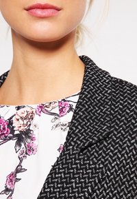 Límec černobíle vzorovaného blejzru překrývá květinovou blůzu s růžovými a fialovými detaily. Hladký povrch, ostré klopy, elegantní střih.