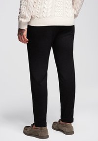 Personne portant un pantalon noir, un pull en laine beige et des mocassins en daim marron, se tenant avec la main relâchée sur le côté, un tatouage floral visible au poignet.