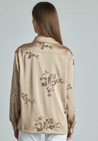 Camicia di satin beige chiaro con ricamo floreale in paillettes. Presenta un colletto, maniche con bottoni e una texture liscia e lucida.