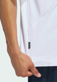 White athletic shirt with a subtle texture, side seam pocket, and a black logo tag on the lower left side. Hand gripping the hem.