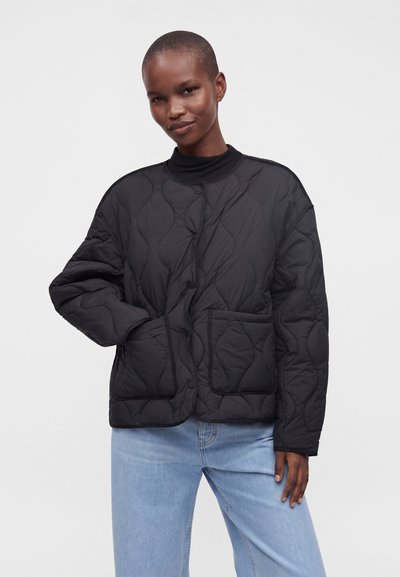 Personne portant une veste matelassée noire avec de grandes poches et un jean bleu clair, debout devant un fond uni et clair.