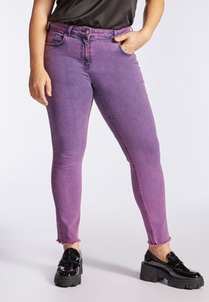 Personne portant un jean violet ajusté avec des ourlets effilochés, un haut noir et des mocassins à plateforme noirs brillants et épais, debout avec une main dans la poche.