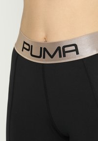Leggings neri con una texture liscia e una vita larga in oro metallizzato, recante il nome del marchio "PUMA" in grassetto nero.
