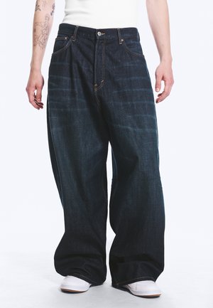 Personne portant un jean large bleu foncé et des baskets blanches, debout devant un fond uni.