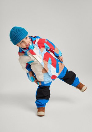 Bunte Regenjacke mit roten, blauen und beigen Mustern; kombiniert mit blauer Strickmütze und schwarzen wasserdichten Hosen; braune Gummistiefel.