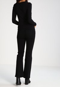 Černý dlouhý rukáv top a černé rozšiřující se kalhoty z hladké látky. Kalhoty mají zadní kapsy; model má na sobě černé boty na podpatku.